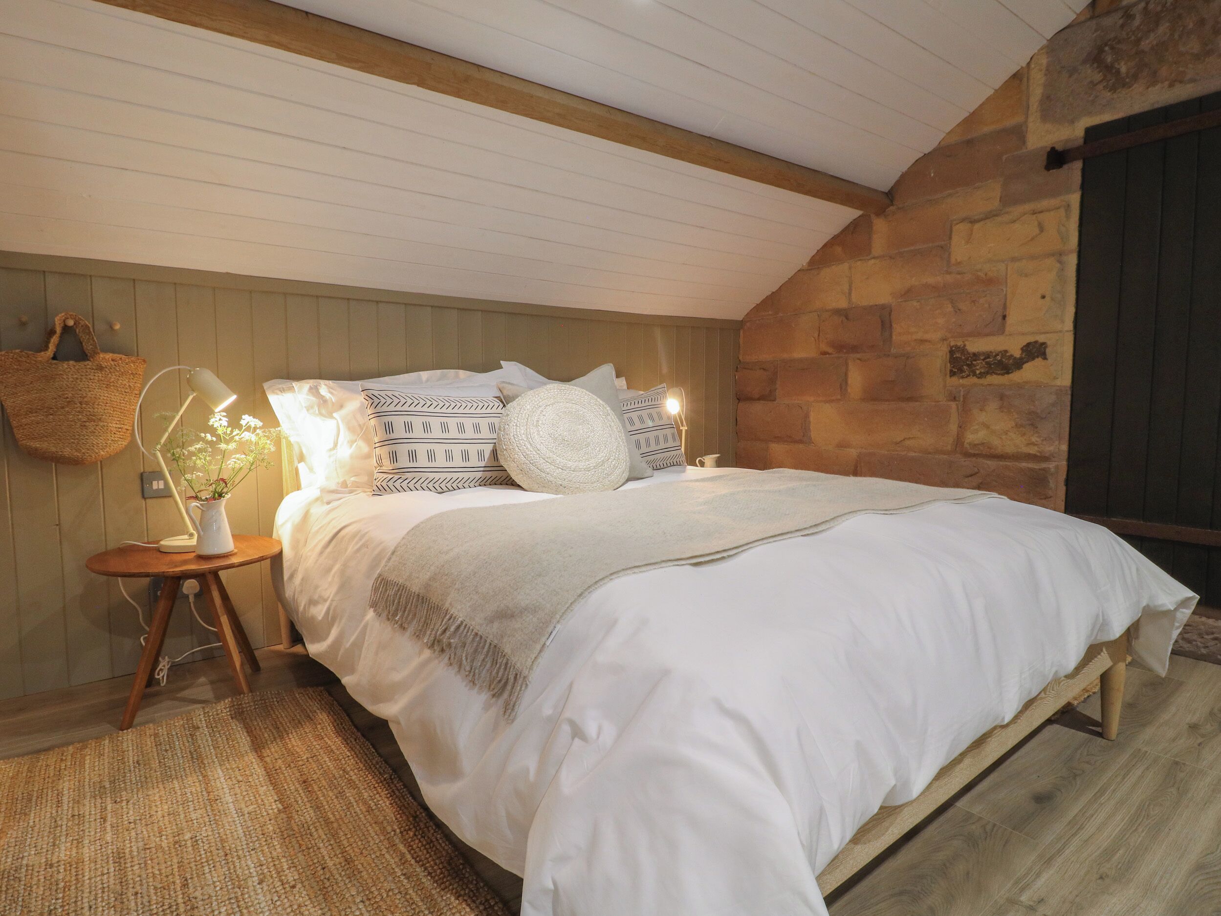 A bedroom with a stone wall, a wooden ceiling, a bed with white bedding, a bedside lamp, and a woven basket on the wall.