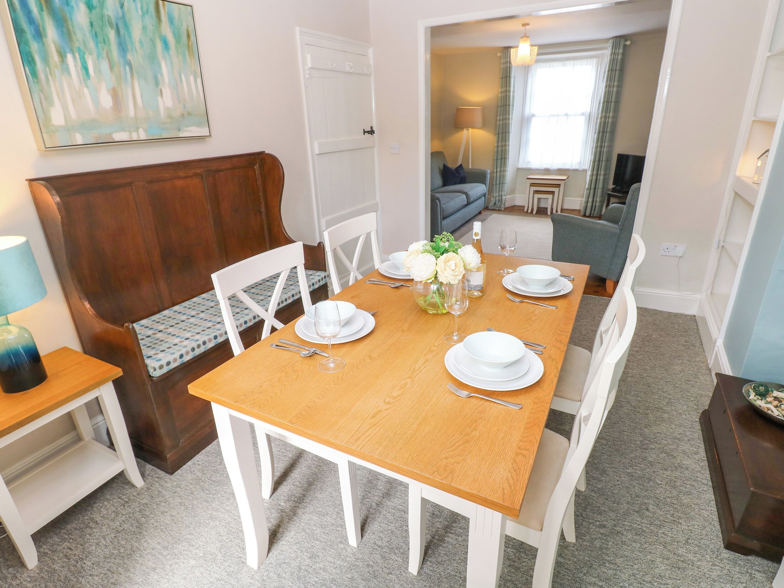 A dining area with a wooden table set for four, white chairs, a bench, and a vase with flowers.