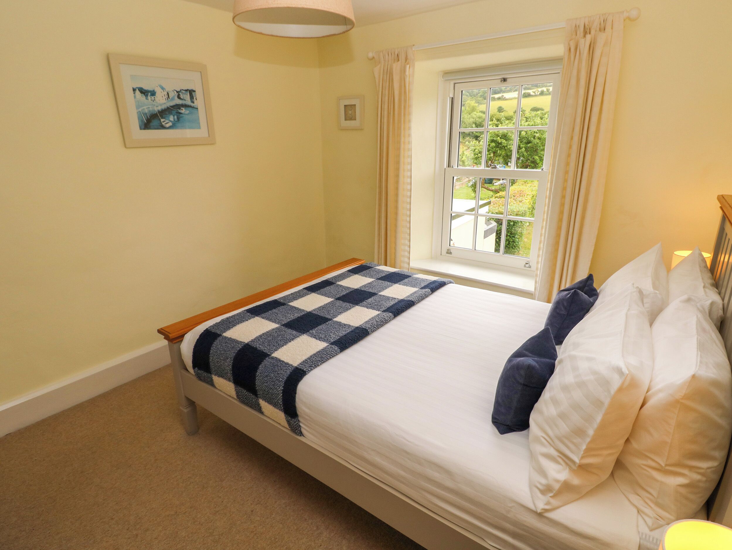 A neatly made bed with a checkered blanket, a window with curtains, and a framed picture on the wall.