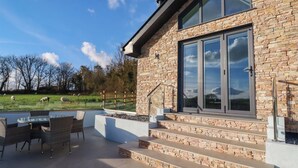 Une maison moderne avec une façade en pierre, de grandes portes vitrées et un patio aménagé avec du mobilier d’extérieur.