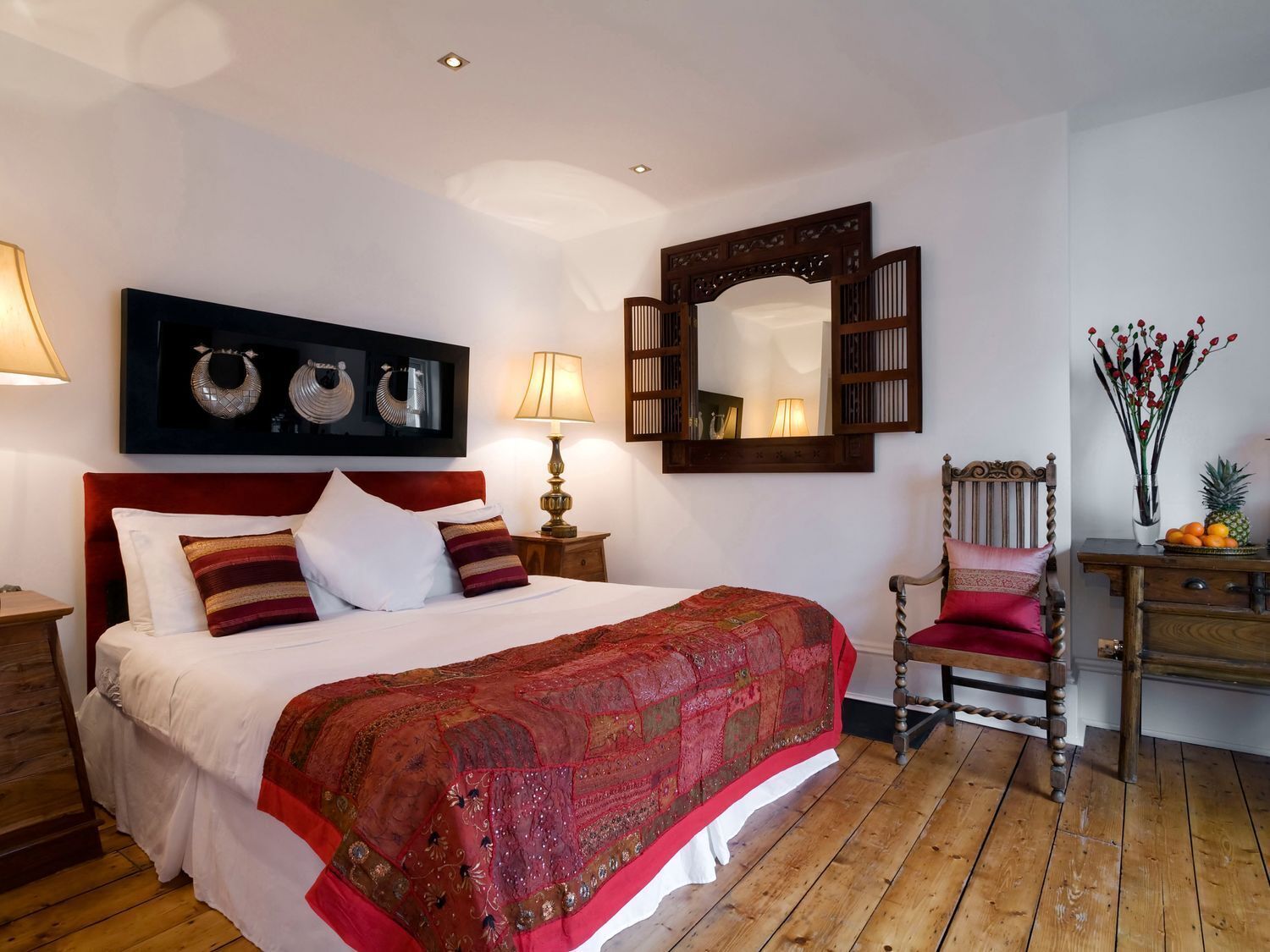 A bedroom with a large bed, a wooden chair, a wooden dresser, a mirror, and wall-mounted artwork.