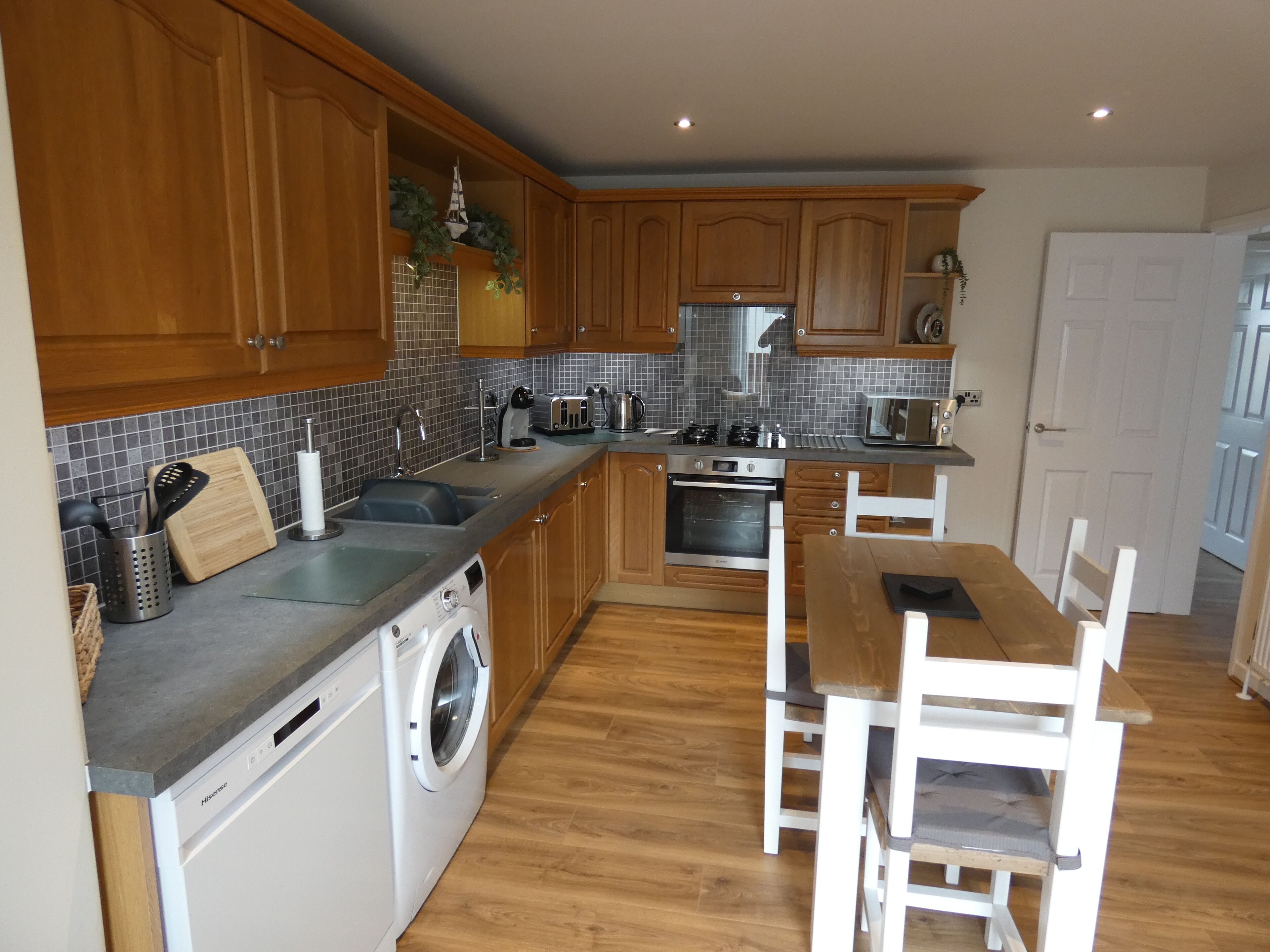 A modern kitchen with wooden cabinets, a built-in oven, a washing machine, and a dining area with a table and chairs.