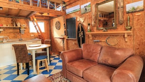 A cozy cabin interior with a wooden ceiling, a dining area with a table and chairs, a leather sofa, and various wall decorations including a mirror and framed pictures. - The Games Room (Chichester)
