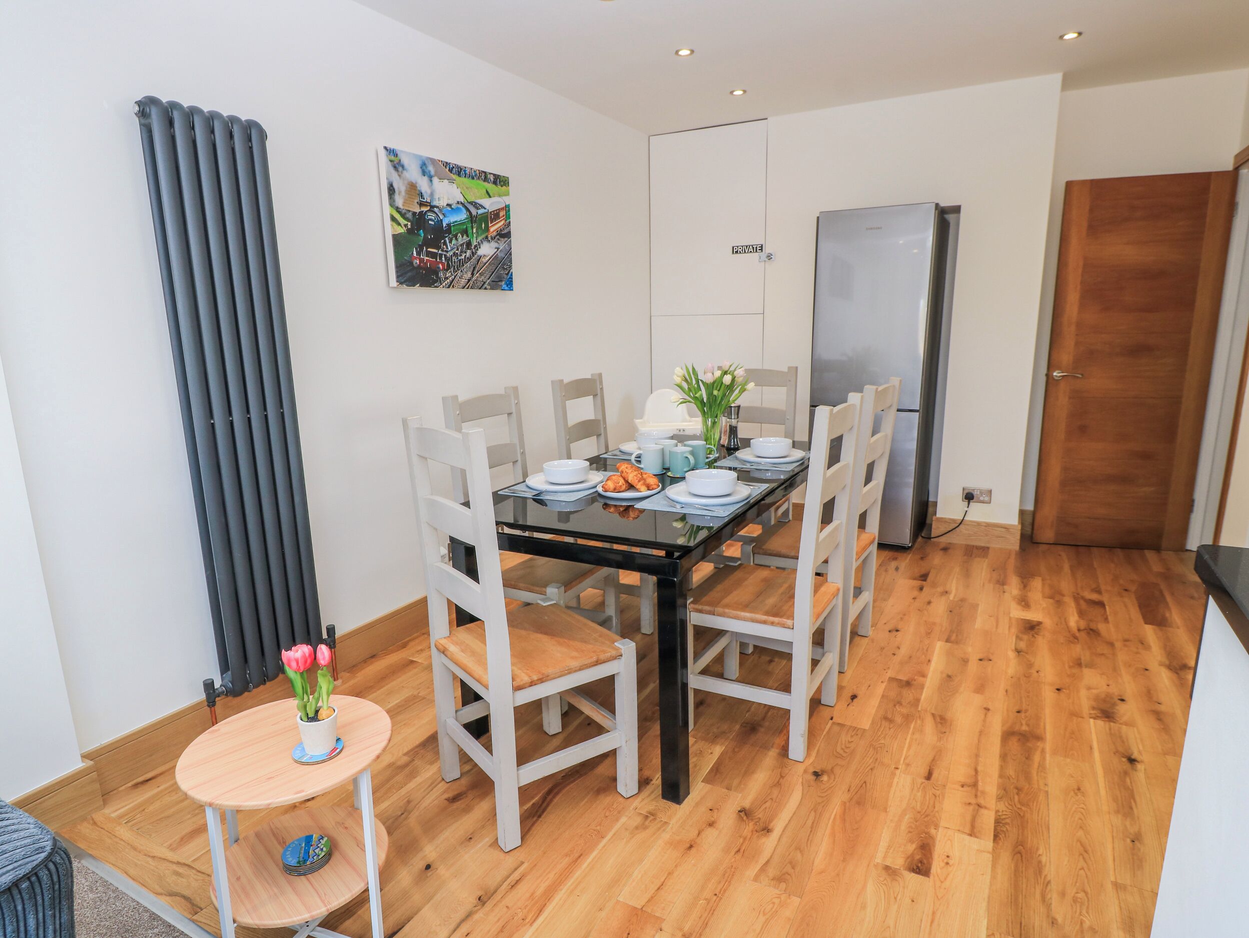 Un comedor con una mesa lista para cuatro personas, sillas, una mesita auxiliar con un jarrón y un refrigerador al fondo.