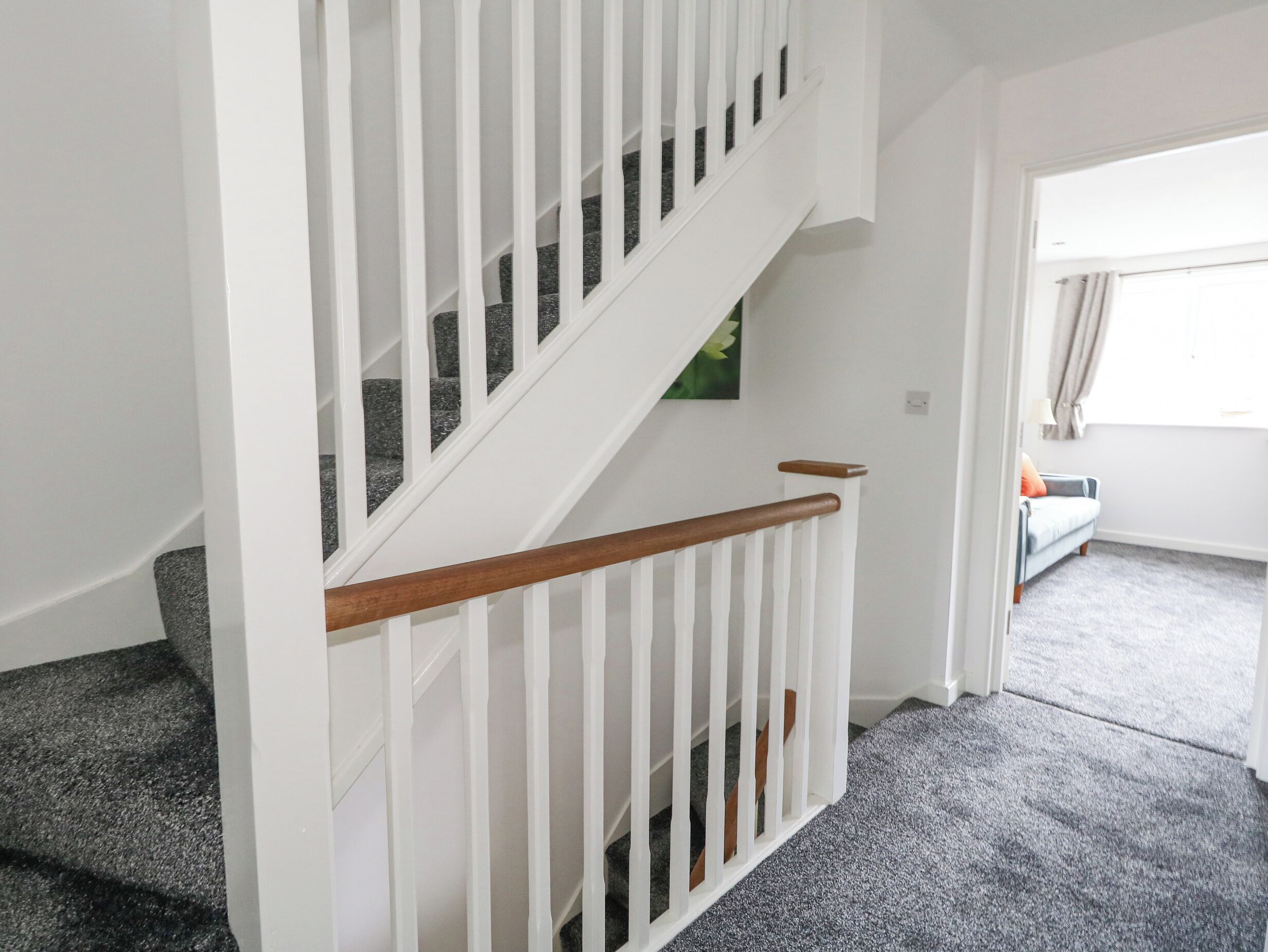 A staircase with white railings and wooden handrails, carpeted steps, and a view of a room with a window and a sofa.