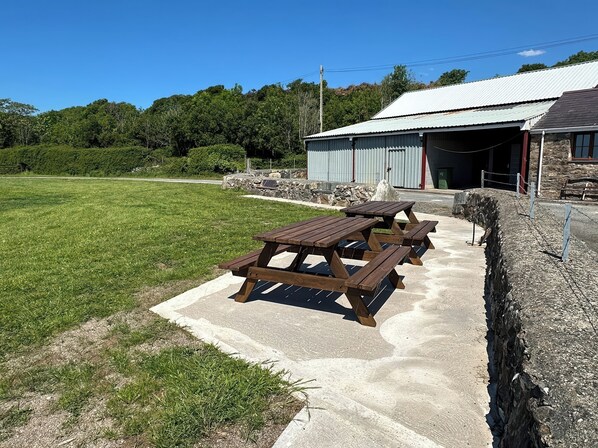 Un espace pique-nique avec des tables et des bancs en bois, un mur en pierre et, au fond, un bâtiment doté d’un toit en métal.
