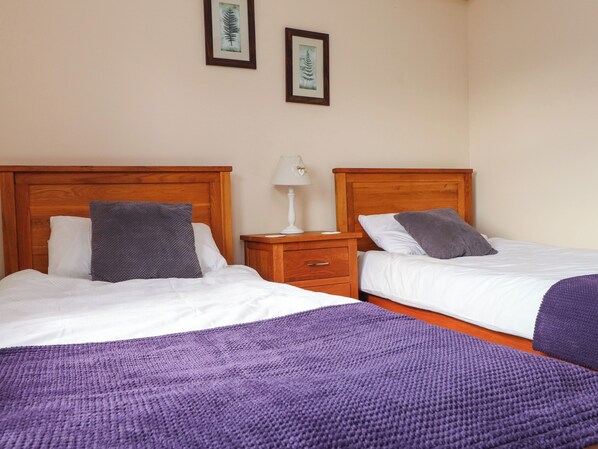 A room with two beds, each with a wooden headboard, white bedding, and a purple throw blanket. There is a nightstand with a lamp and a drawer between the beds. - The Mill (Pwllheli)