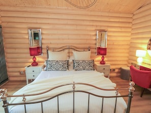 A wooden cabin bedroom with a bed, two bedside tables, two lamps, a mirror, and a red chair. - Carlton Lodge 1 (Middlesbrough)