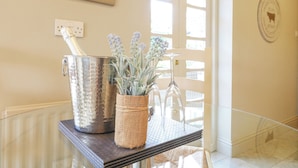Una mesa de vidrio con un recipiente metálico y una cesta tejida que contiene lavanda. Se ve una puerta con un panel de vidrio y un emblema fijado en la pared.