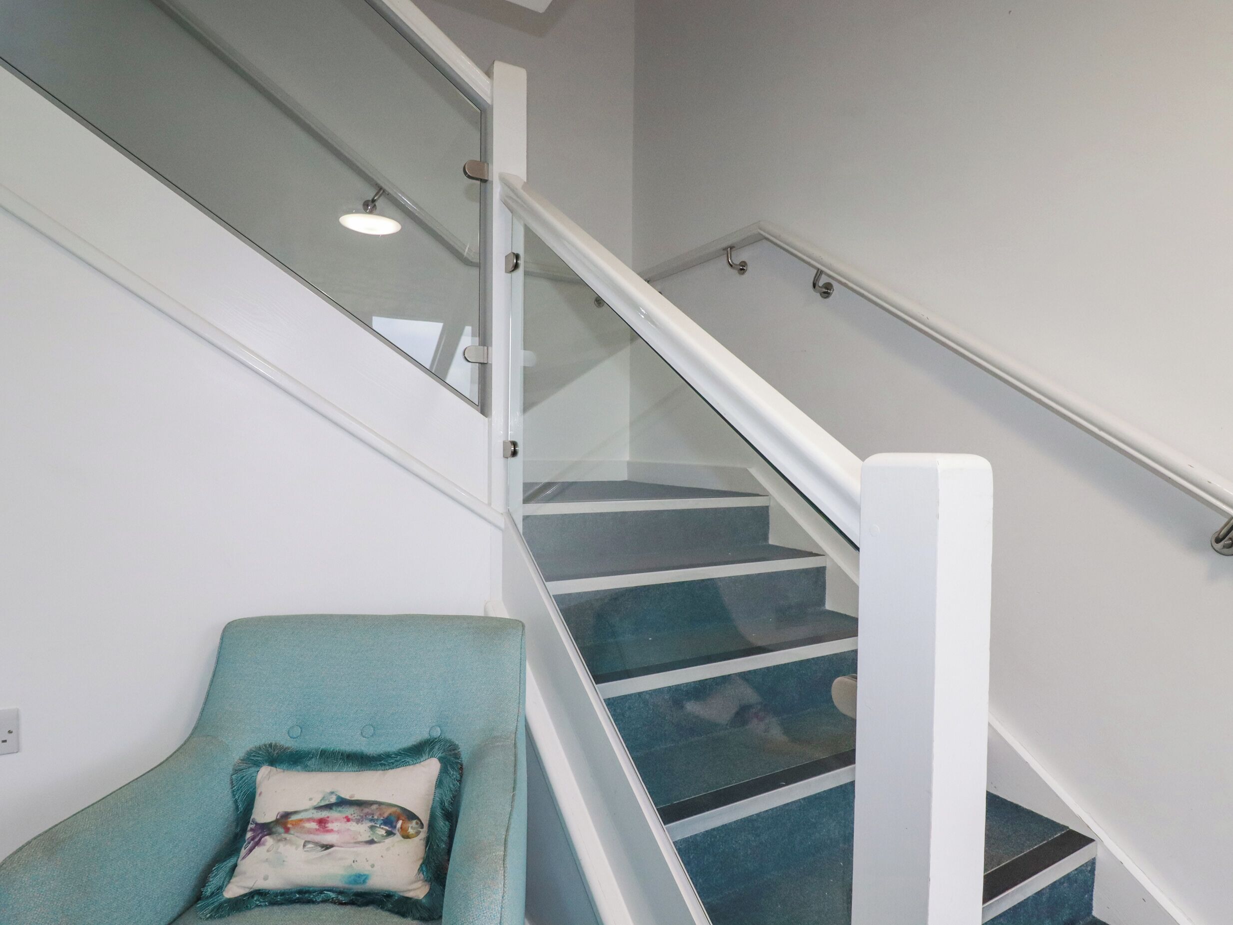 Un escalier avec des rampes en verre et un fauteuil turquoise agrémenté d’un coussin décoratif.