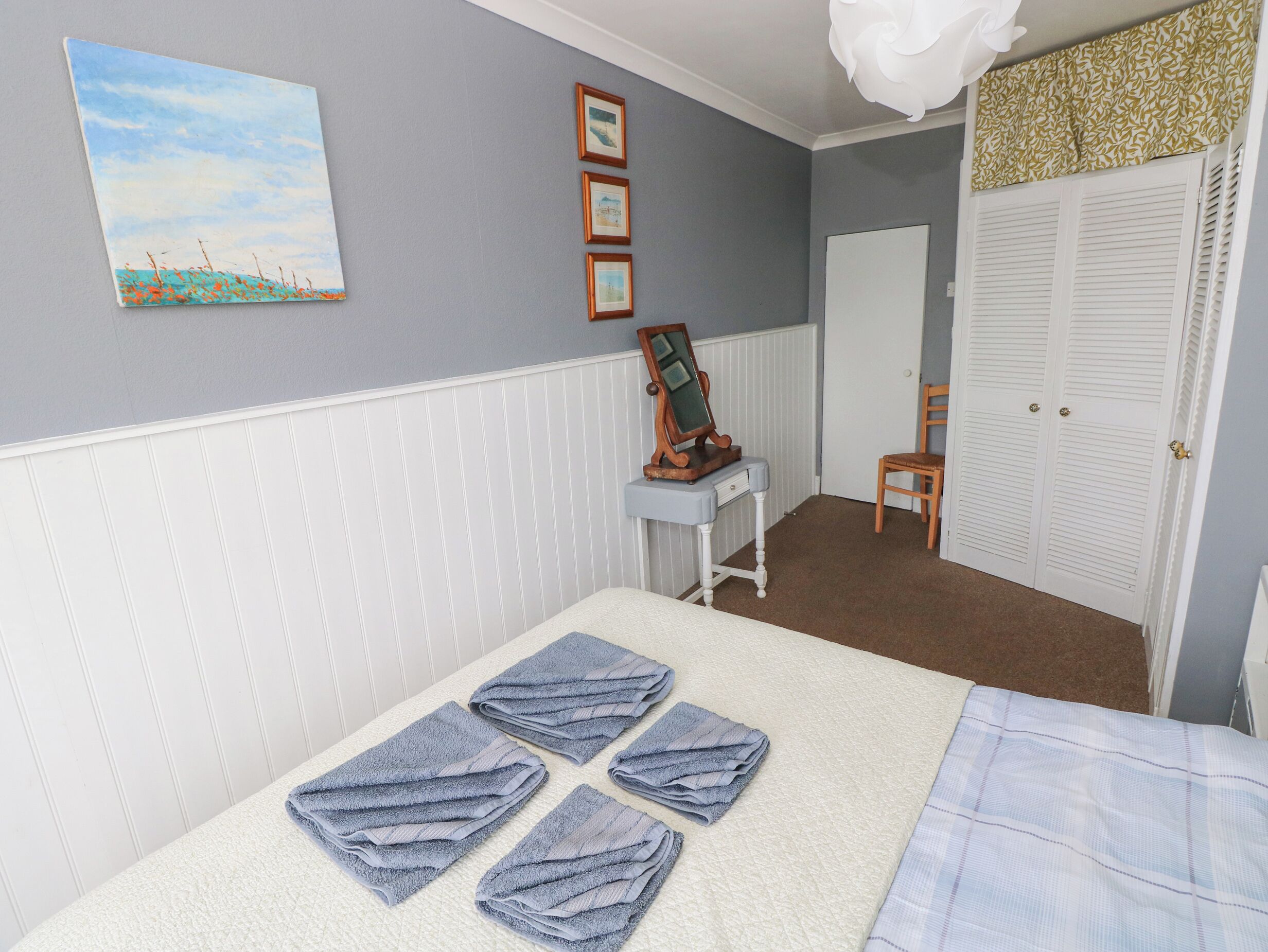 Une chambre à coucher bien rangée, avec un lit, une coiffeuse dotée d’un miroir, une chaise et une porte munie de volets à persiennes.