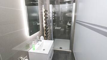 Moderni kylpyhuone, jossa on lasinen suihkuseinä, valkoinen pesuallas ja sen yläpuolella peili sekä wc-istuin.