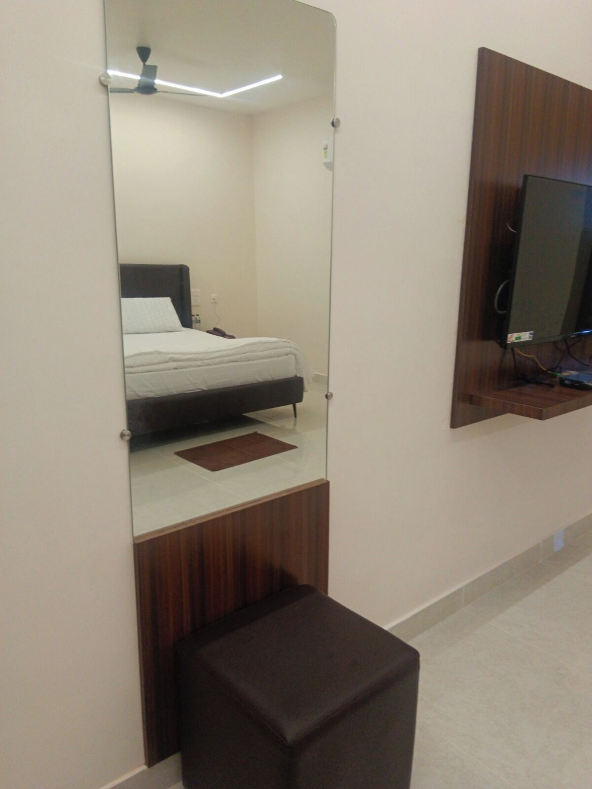 Un dormitorio con cama, mesita de noche, espejo, televisor montado en la pared y un pequeño banco de madera.