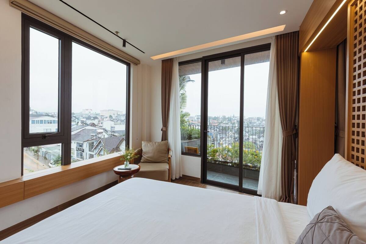 Habitación de hotel con una cama grande, una silla, una mesita con un florero y un balcón con vistas a la ciudad.
