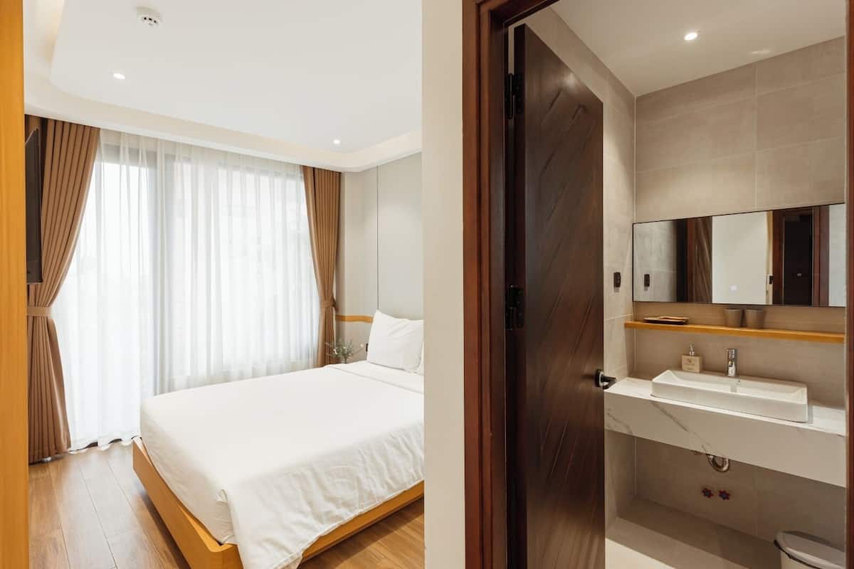 Habitación de hotel con cama, cortinas, suelo de madera y baño con lavamanos y espejo.
