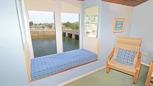 A room with a large window overlooking a bridge and water, a wooden chair with a cushion, and a framed picture on the wall. - Glaslyn Retreat (Porthmadog)