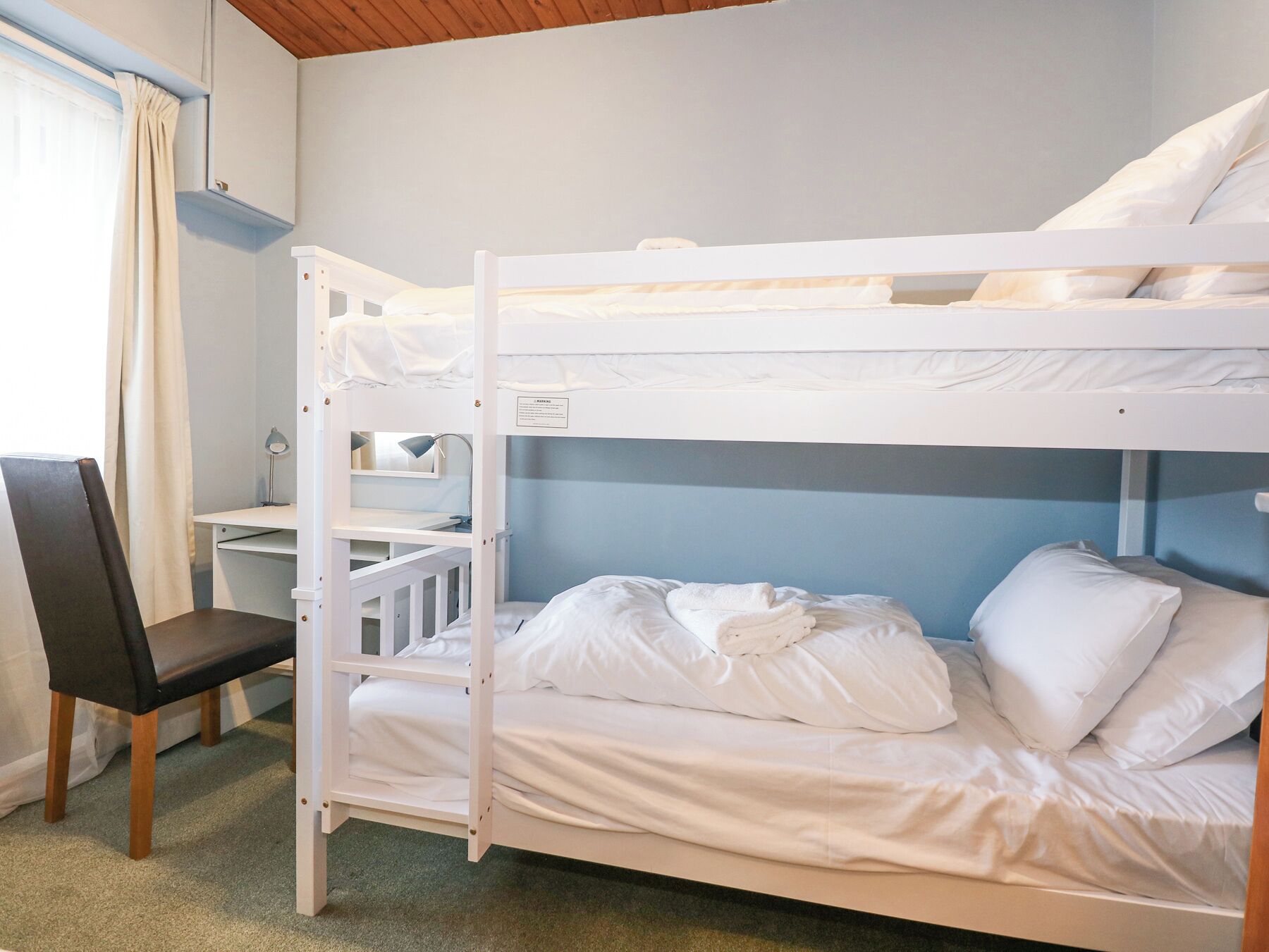 Une chambre avec un lit superposé, un bureau et une chaise, une fenêtre avec des rideaux et un plafond en bois.