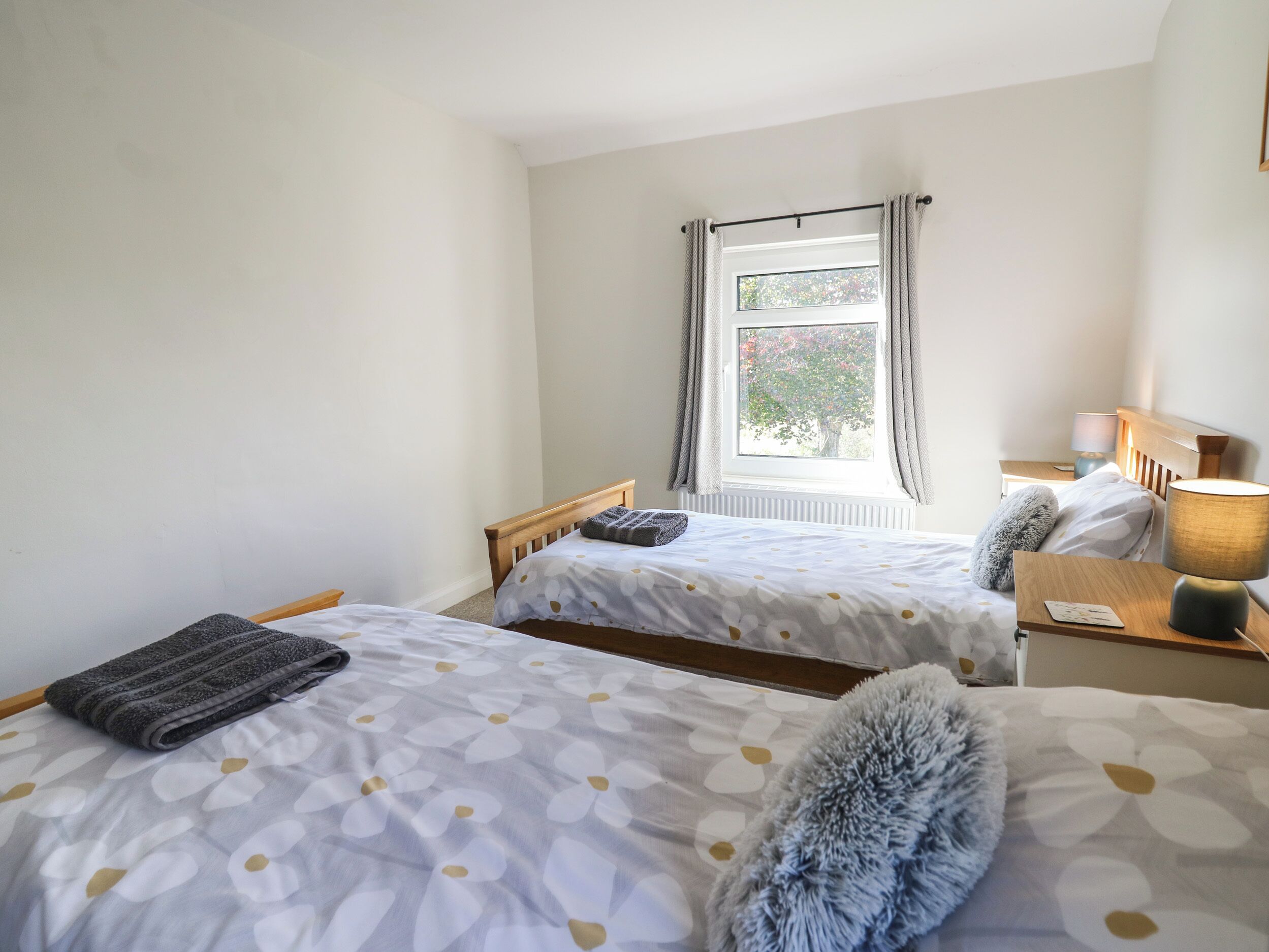 A bedroom with two beds, a window with curtains, a bedside table with a lamp, and a radiator.