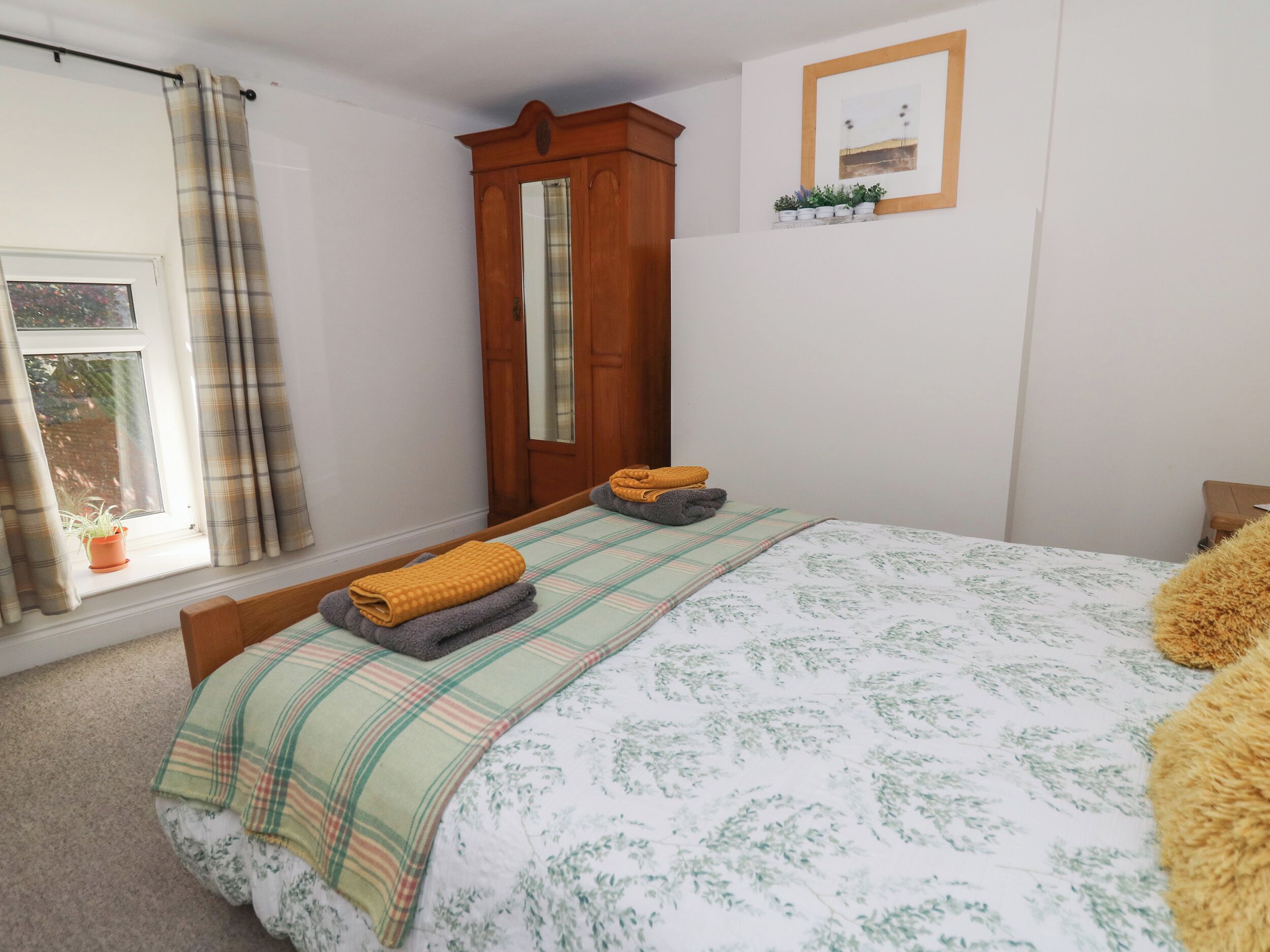 A bedroom with a bed, a wardrobe, a mirror, a window with curtains, and a small shelf with plants.