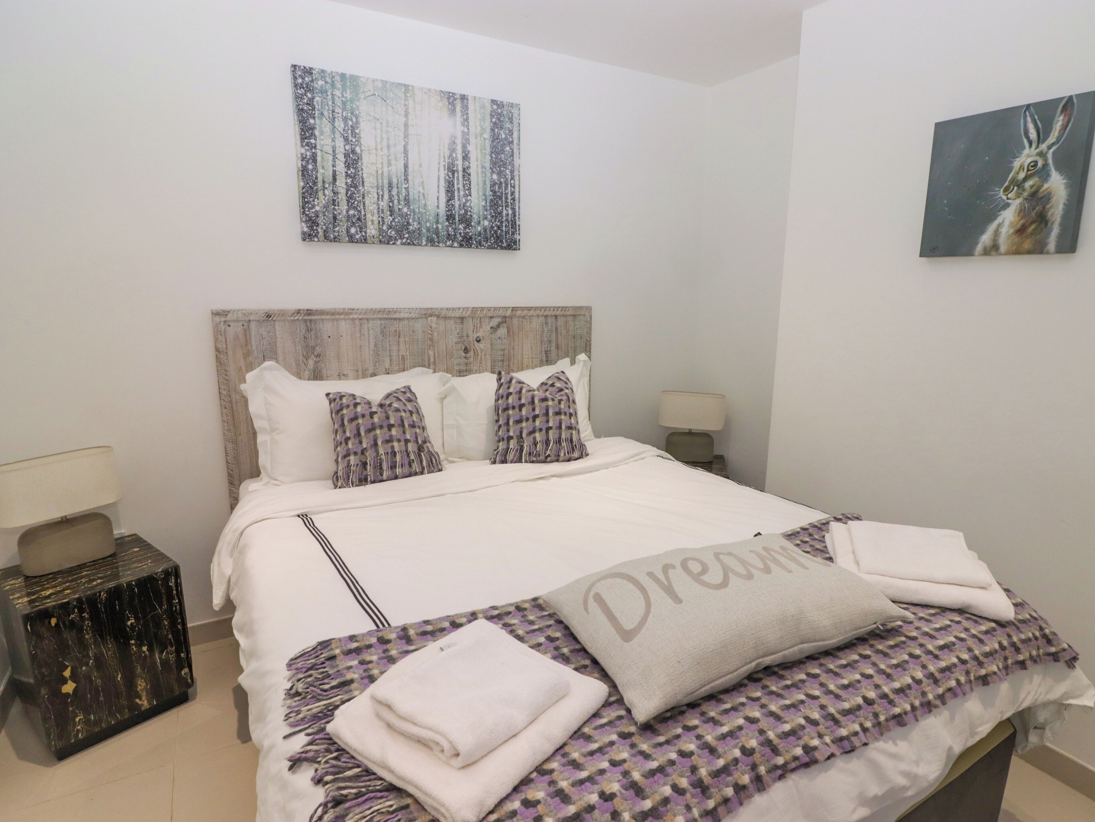 Una cama bien hecha con un edredón estampado, una almohada con la palabra 'Dream' y un cuadro de un conejo en la pared.