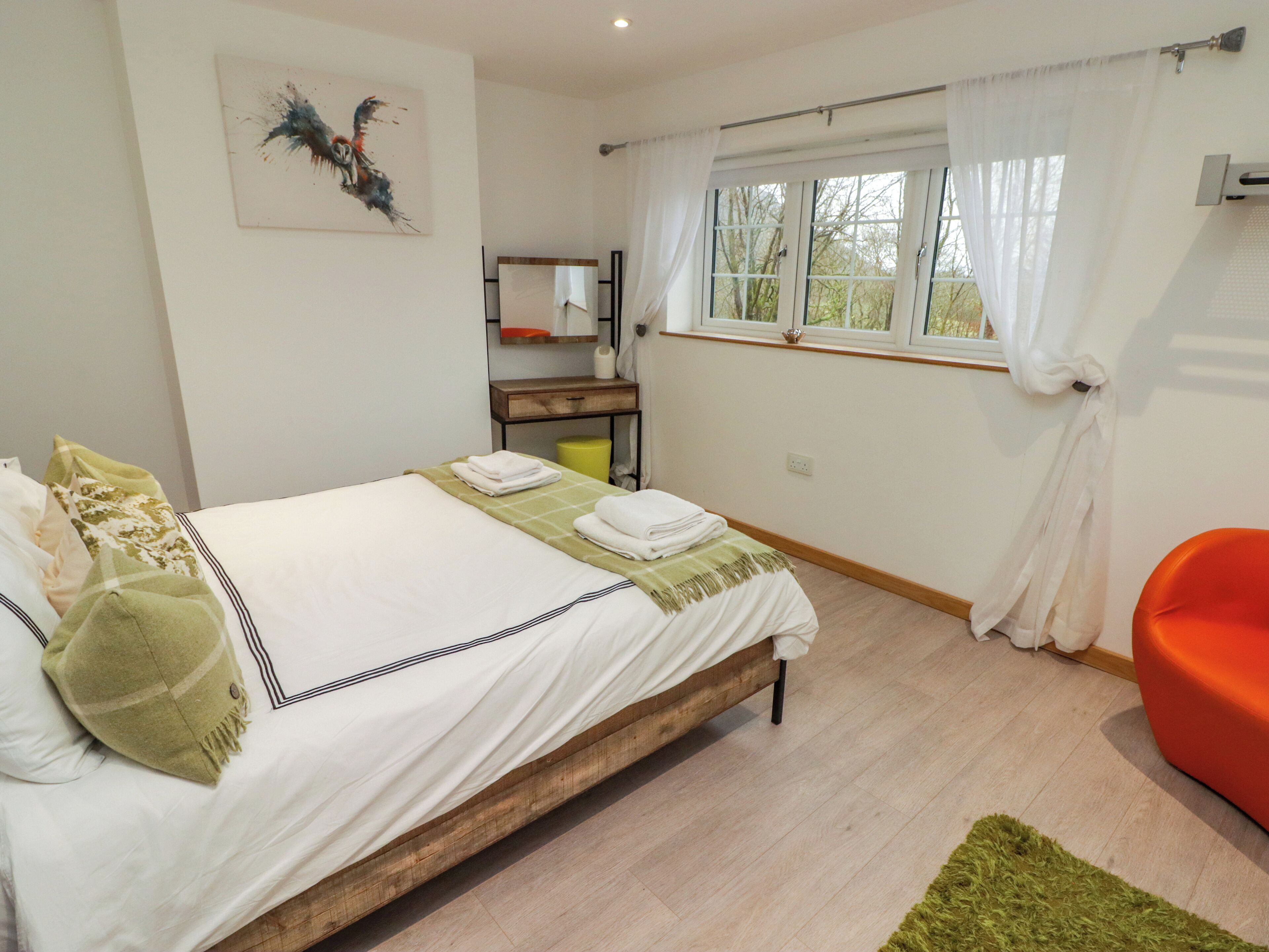 Een slaapkamer met een bed, nachtkastje, stoel, raam met gordijnen en een schilderij aan de muur.
