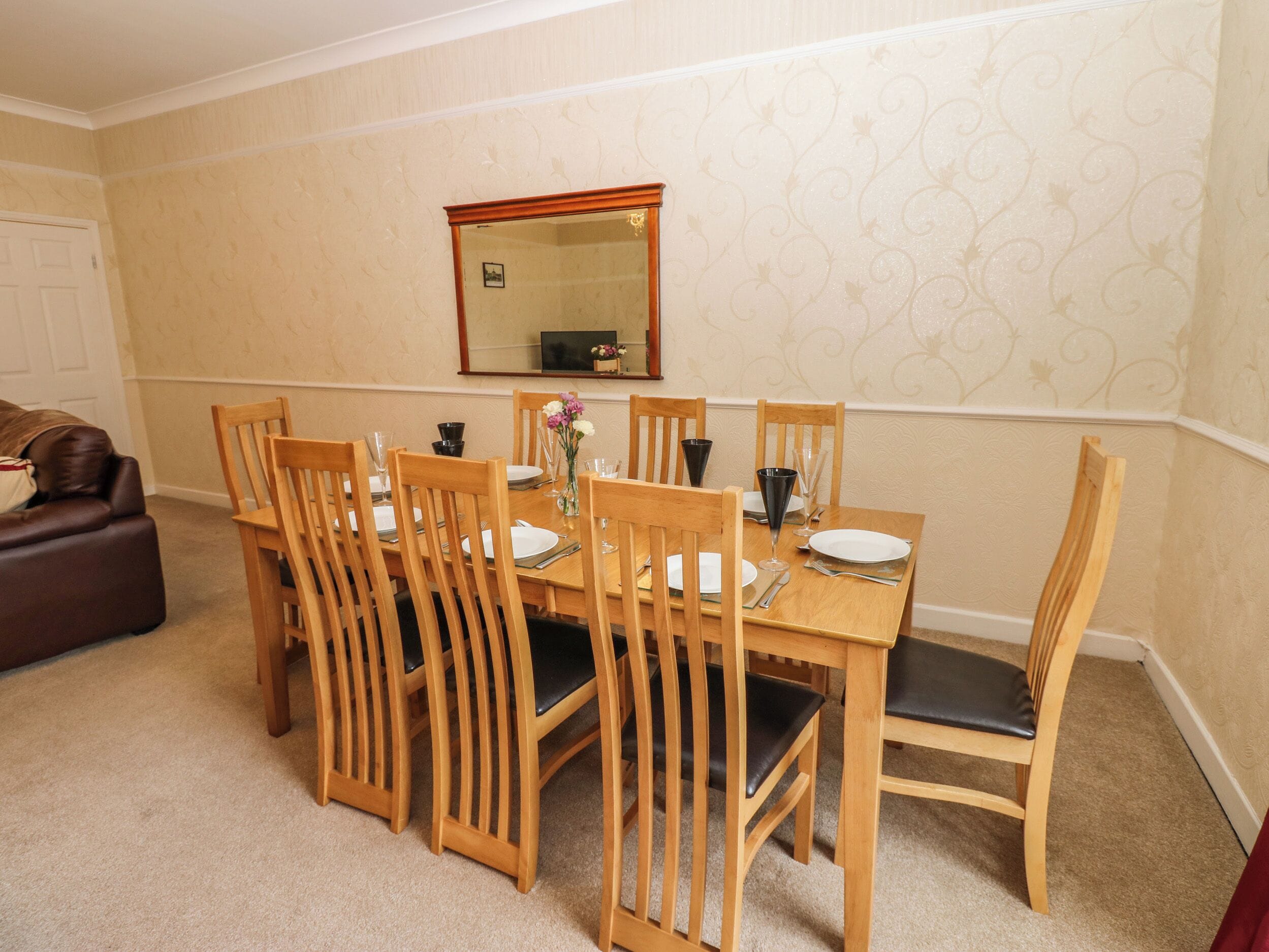 A dining area with a wooden table set for six, surrounded by six wooden chairs with black cushions.