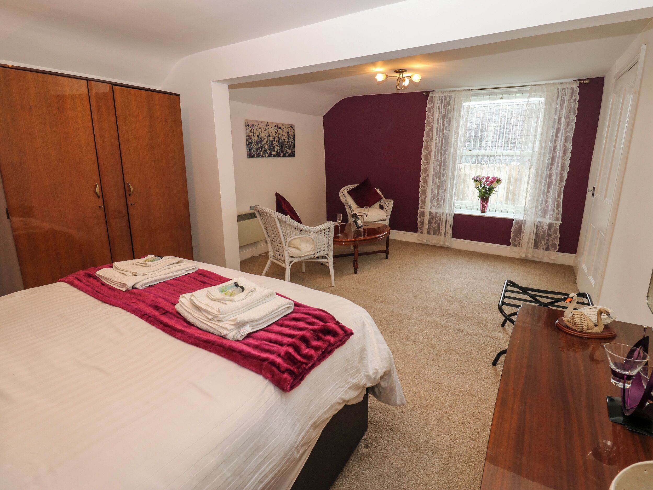 A bedroom with a bed, a wooden wardrobe, a wicker chair, a small table, and a window with curtains.