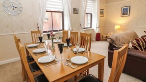 A dining area with a wooden table set for six, surrounded by chairs. There is a clock on the wall, a window with curtains, and a sofa in the background. - The Old Assembly Rooms (Bishop Auckland)
