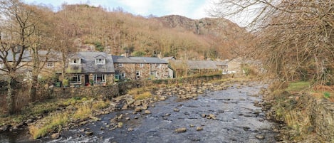 A stone building with a pub sign, a river with rocks, and a hillside in the background.