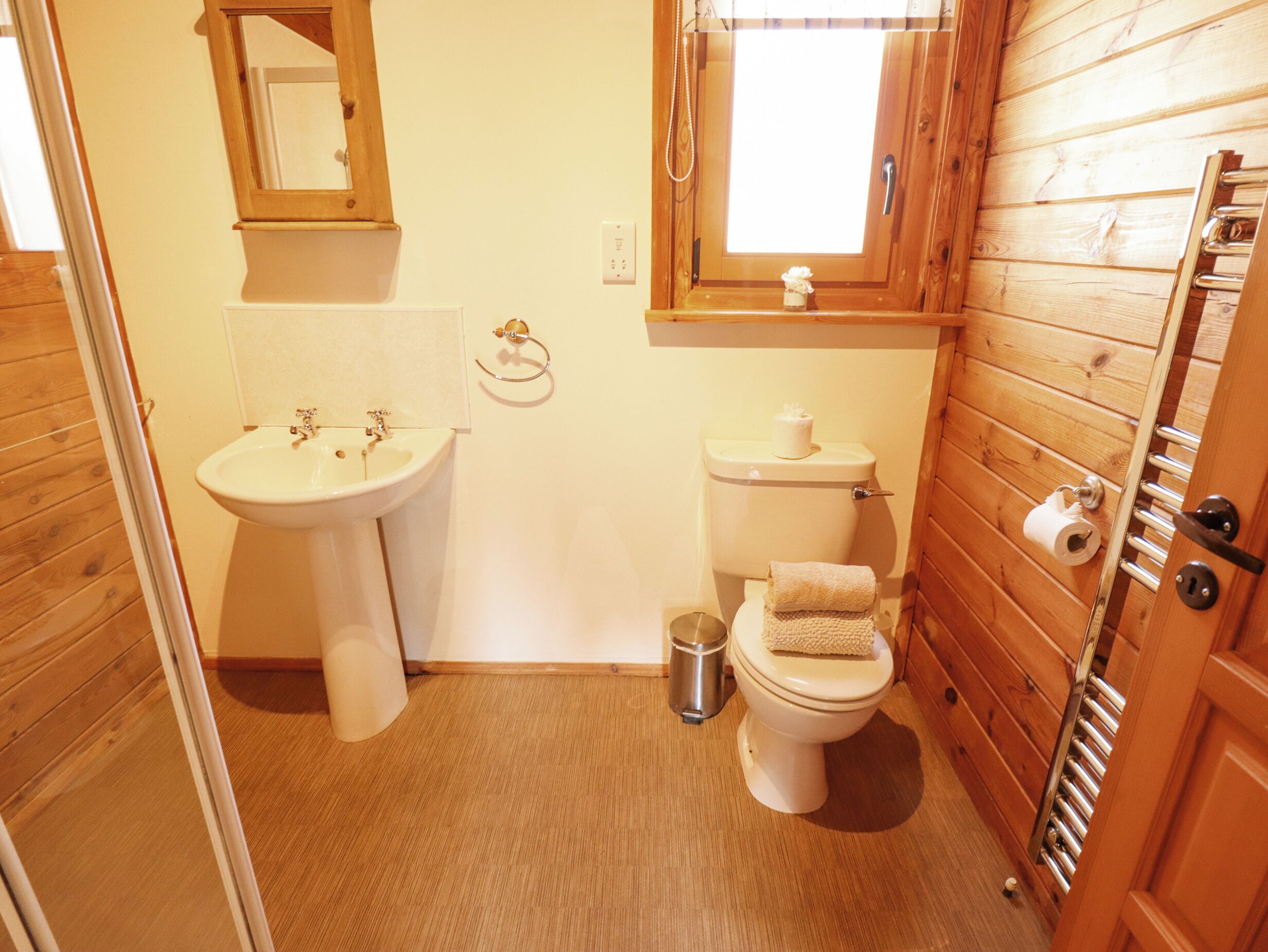 A small bathroom with a wooden floor, a white sink, a toilet, and a window above the toilet.