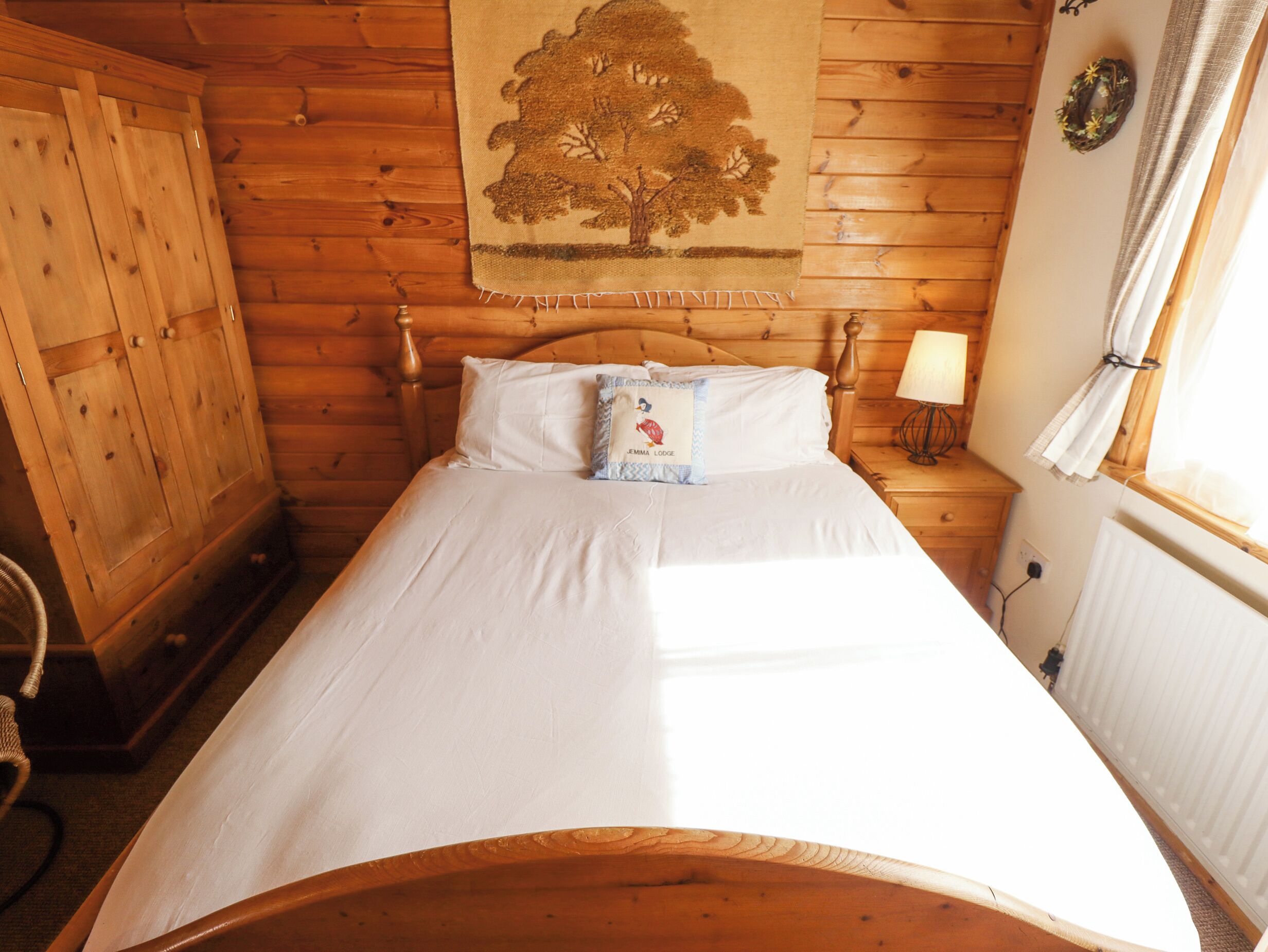 Habitación de cabaña de madera con cama, mesita de noche con lámpara, ventana con cortinas y armario de madera.