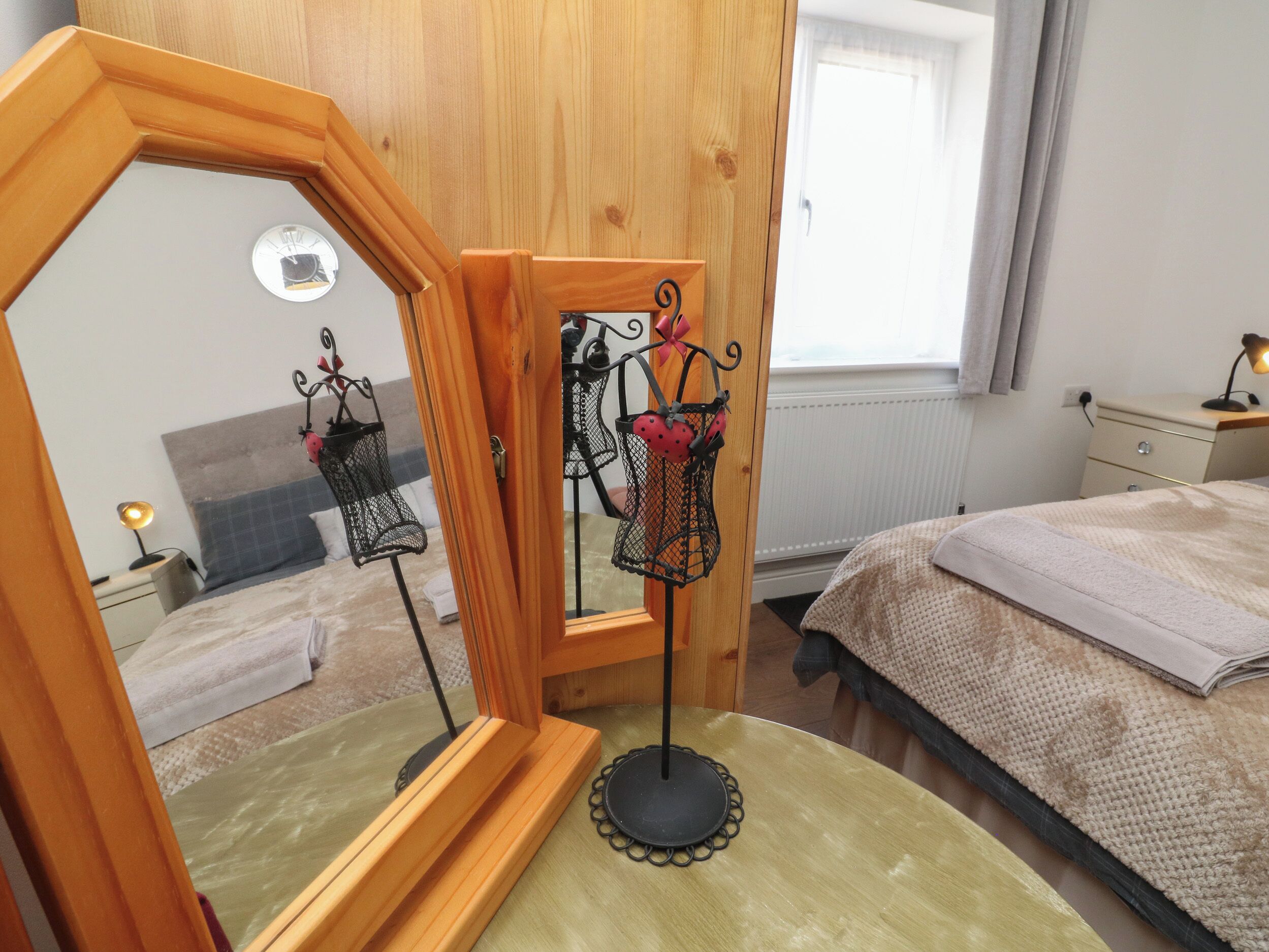 A bedroom with a wooden wardrobe, a mirror, a bedside table with a lamp, a bed with a beige blanket, and a window with curtains.