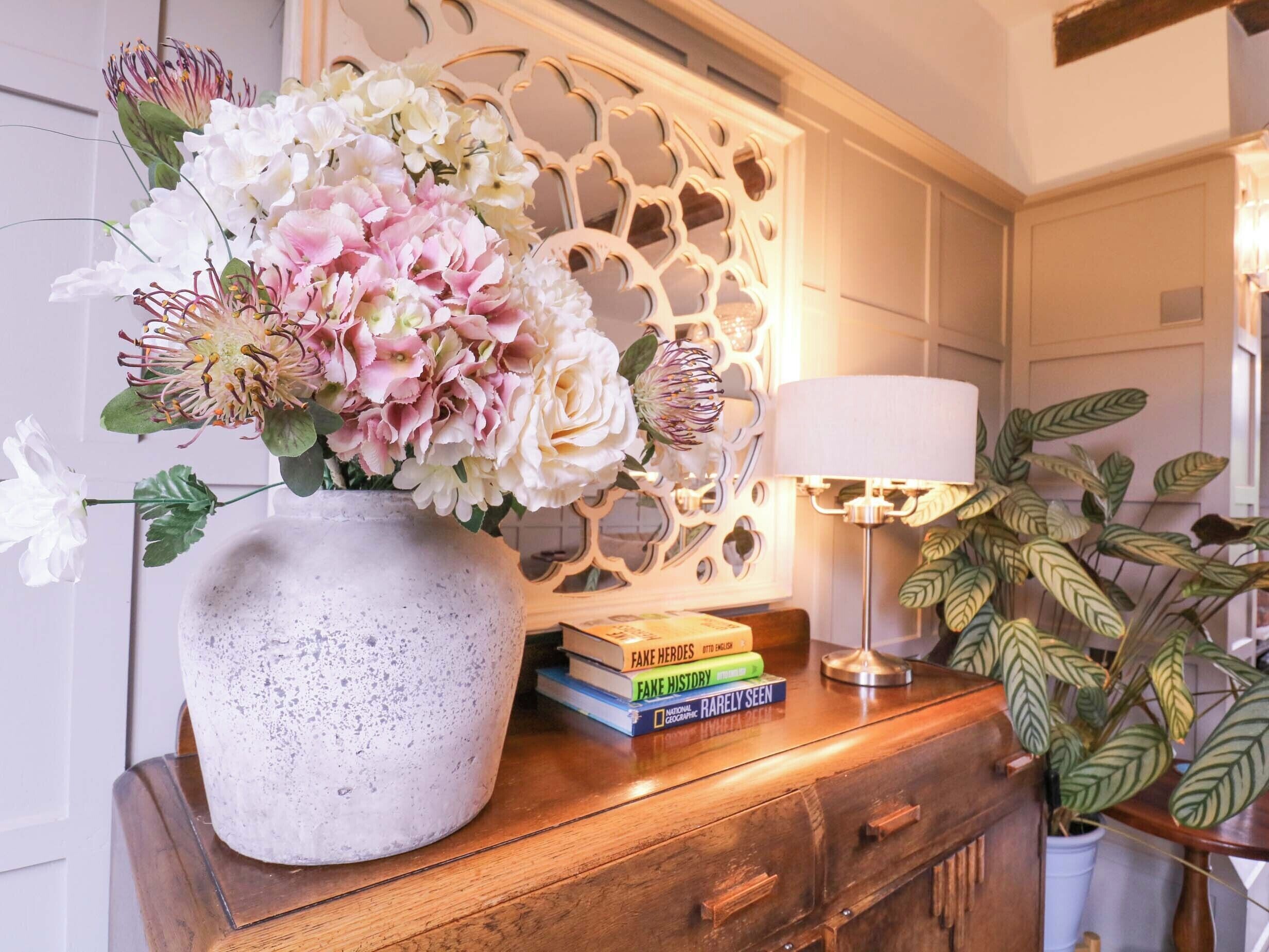 Un arreglo floral en una maceta moteada sobre una mesita de madera, junto a una pila de libros y una lámpara.