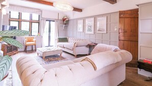 Un salón amplio con un sofá grande, un sillón más pequeño y una mesa de centro. A la derecha hay una puerta de madera y a la izquierda una ventana con cortinas.