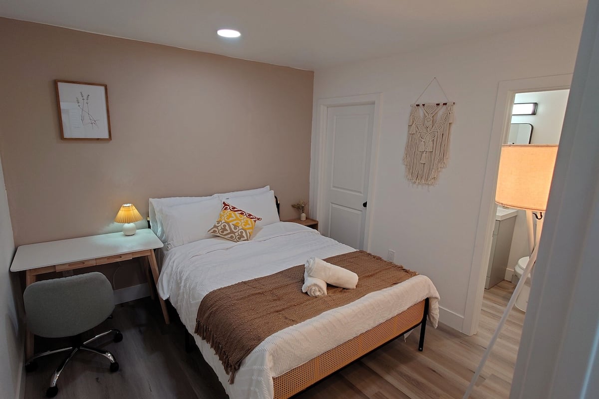 Une chambre à coucher comprenant un lit, un bureau, une chaise, une lampe de chevet et une décoration murale en macramé.