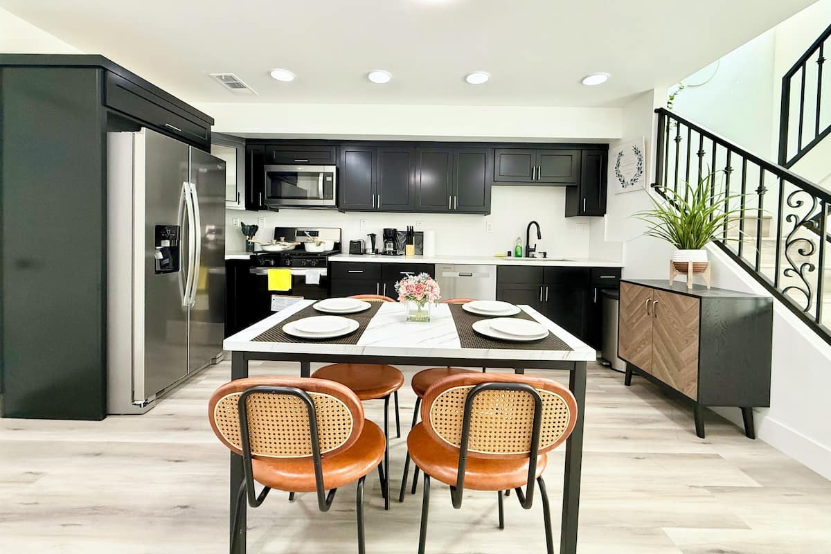 Une cuisine moderne avec des meubles de couleur sombre, des appareils en acier inoxydable et un coin repas comprenant une table blanche et des chaises en osier.
