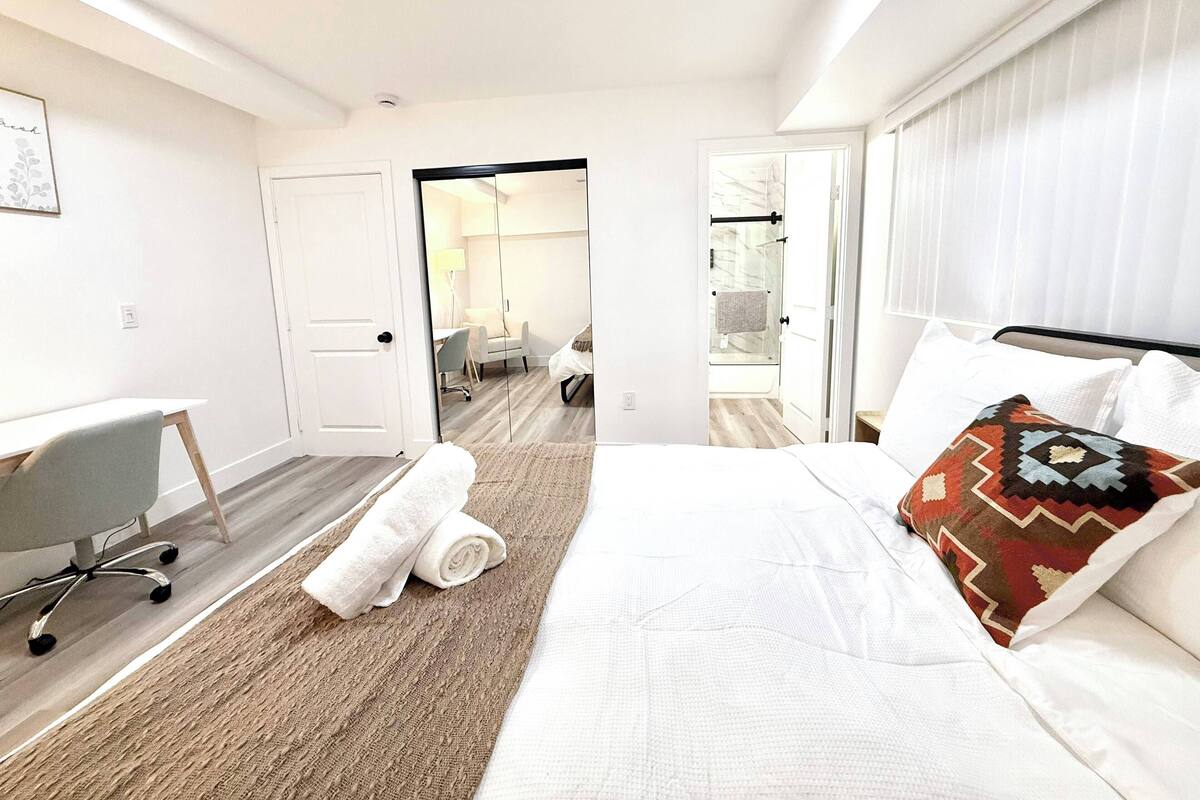 Une chambre d’hôtel avec un lit, un bureau et une chaise. On y trouve une porte donnant sur une salle de bain et un miroir.