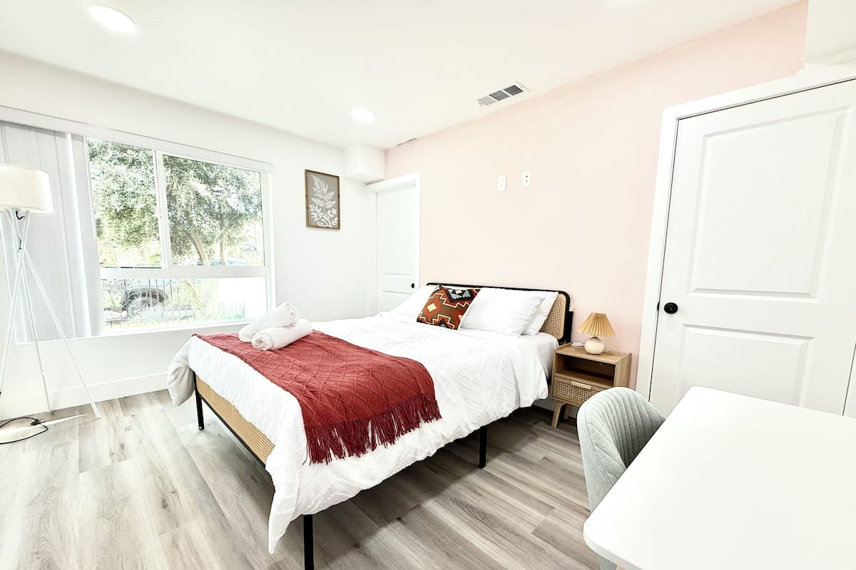 Une chambre à coucher comprenant un lit, une table de chevet, une lampe, une chaise et une fenêtre donnant sur des arbres.