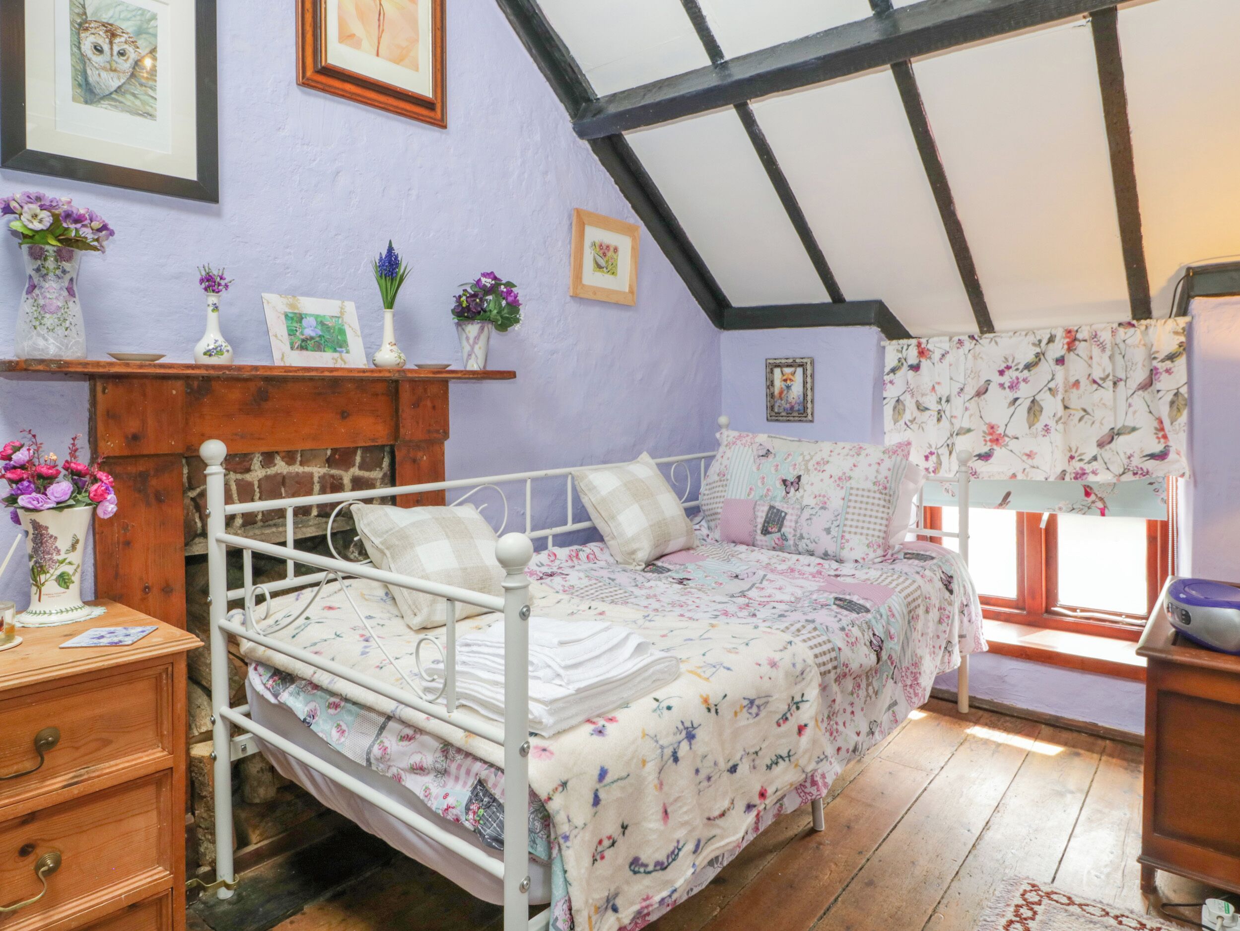 Un dormitorio con techo inclinado, una cama con ropa de cama floral, un aparador y una ventana con cortinas florales.