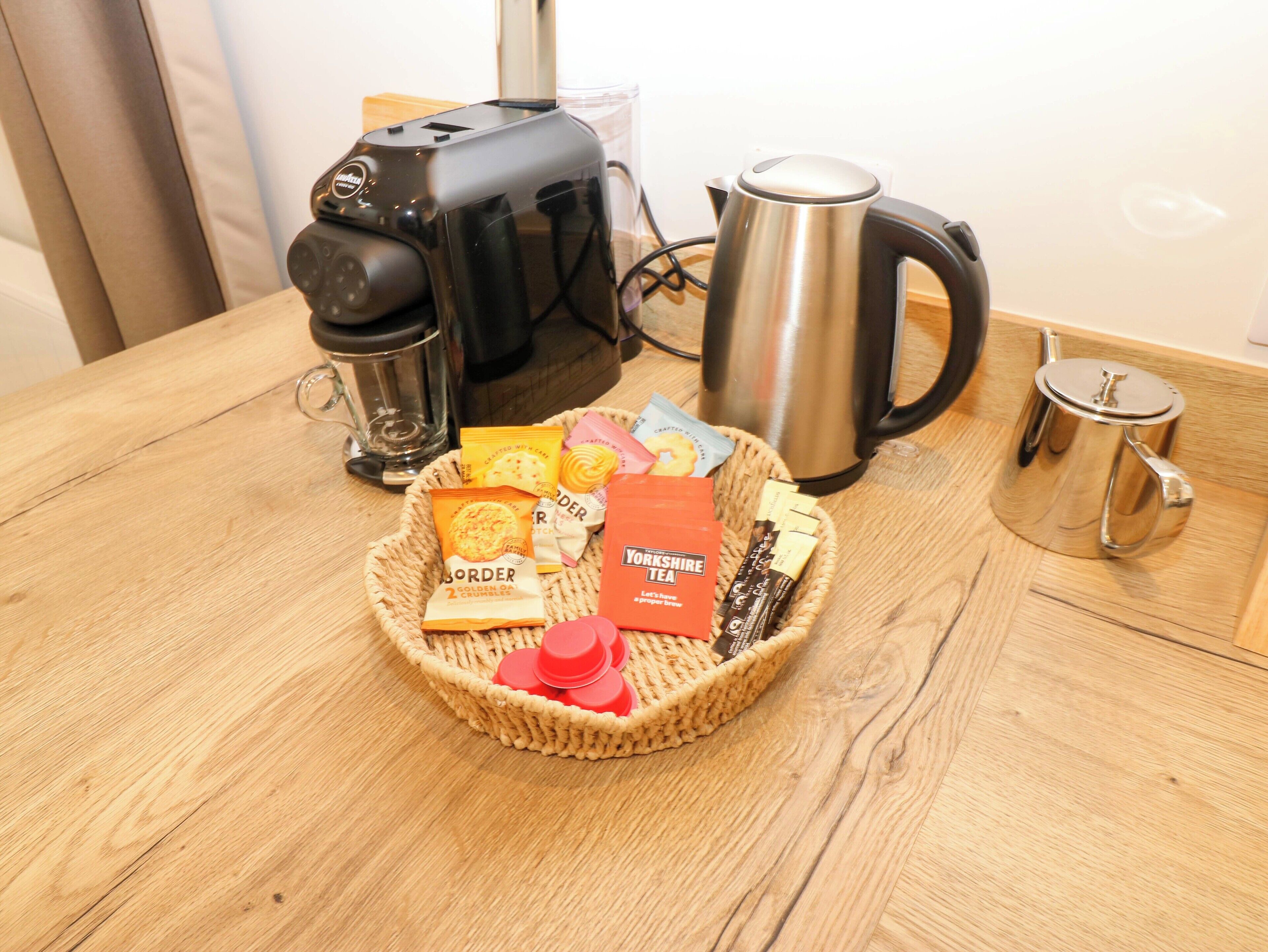 A coffee maker, a kettle, and a basket with tea and snacks on a wooden table.