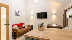 Een moderne woonkamer met een houten eettafel, een bank, een aan de muur bevestigde televisie, en een keuken met een houten aanrecht.