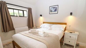 Een net opgemaakt bed met witte lakens en een beige deken, een nachtkastje met een lamp, en een ingelijst beeld aan de muur.