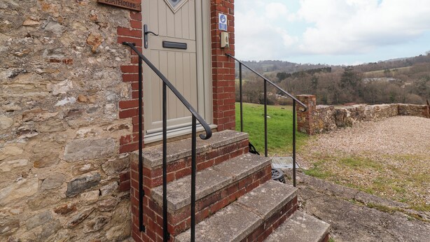 A stone building with a beige door, a metal staircase, and a view of a grassy field. - The Goathouse Cottage (Lyme Regis)