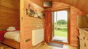 A cozy wooden cabin room with a bed, a mounted TV, and a view of a grassy field through a sliding door. - Oak View (Llangadog)