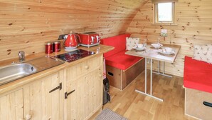A cozy wooden cabin with a kitchen area featuring a red toaster, kettle, and a sink. There is a red sofa and a small table with a white tablecloth, along with a window providing natural light. - Oak View (Llangadog)