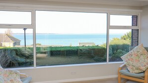 Ein Zimmer mit einem großen Fenster mit Blick auf ein Gewässer, zwei Sessel mit Blumenkissen und einer Leuchte darüber.