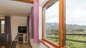 Ein Zimmer mit Holzbalkendecke, einem Fernseher auf einem Holzständer, einem Fenster mit Blick auf eine ländliche Landschaft und violetten Vorhängen.