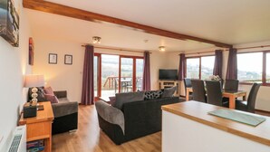 Ein großzügiger Wohnbereich mit Holzboden, einer Couch, einem Couchtisch und einem Fernseher. Große Fenster mit Vorhängen, ein Balkon und ein Blick ins Freie sind vorhanden.