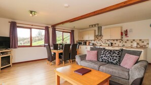 Ein gemütliches Wohnzimmer mit einem Couchtisch aus Holz, einer Couch mit gemusterten Kissen und einem Essbereich mit Tisch und Stühlen. Im Hintergrund ist eine Küche mit Holzschränken und einer gefliesten Rückwand zu sehen.