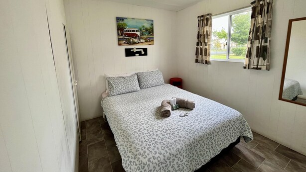 A bedroom with a bed, a window with curtains, a painting on the wall, and a small red stool. - Cootamundra Caravan Park (Cootamundra)